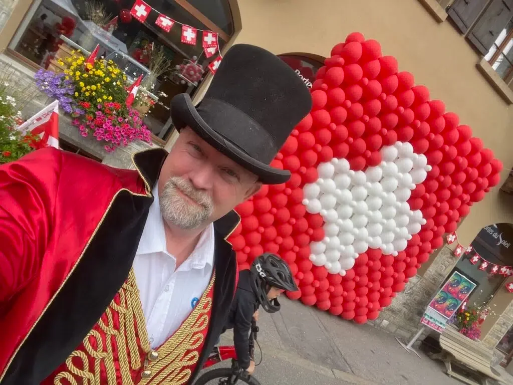 Tampilipe con cilindro e giacca rossa da direttore di circo davanti a un cuore di palloncini con la bandiera svizzera